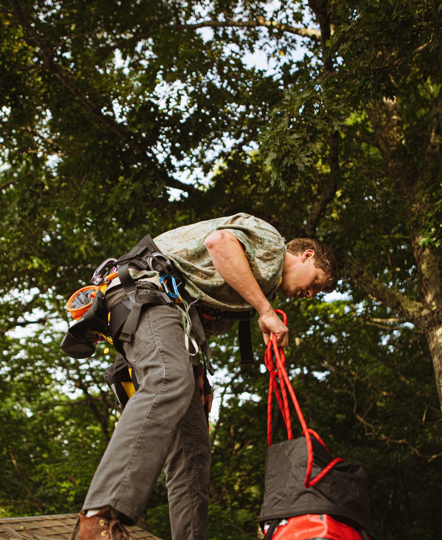 Roofer securing equipment on a shingled roof among trees